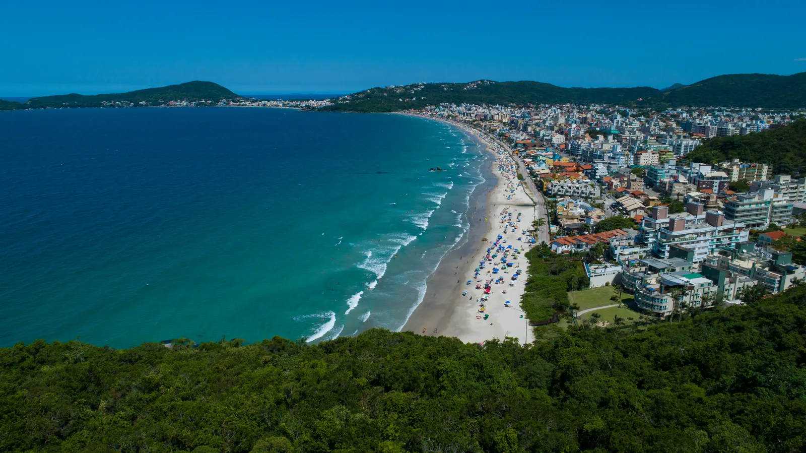 Vista aérea da Praia de Bombas: faixa de areia curva, mar turquesa e o morro coberto de mata contornando a orla