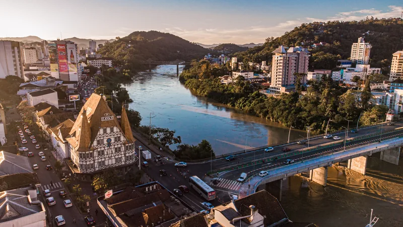 Vista aérea de Blumenau ao entardecer, com o rio cortando a cidade e a ponte ao fundo