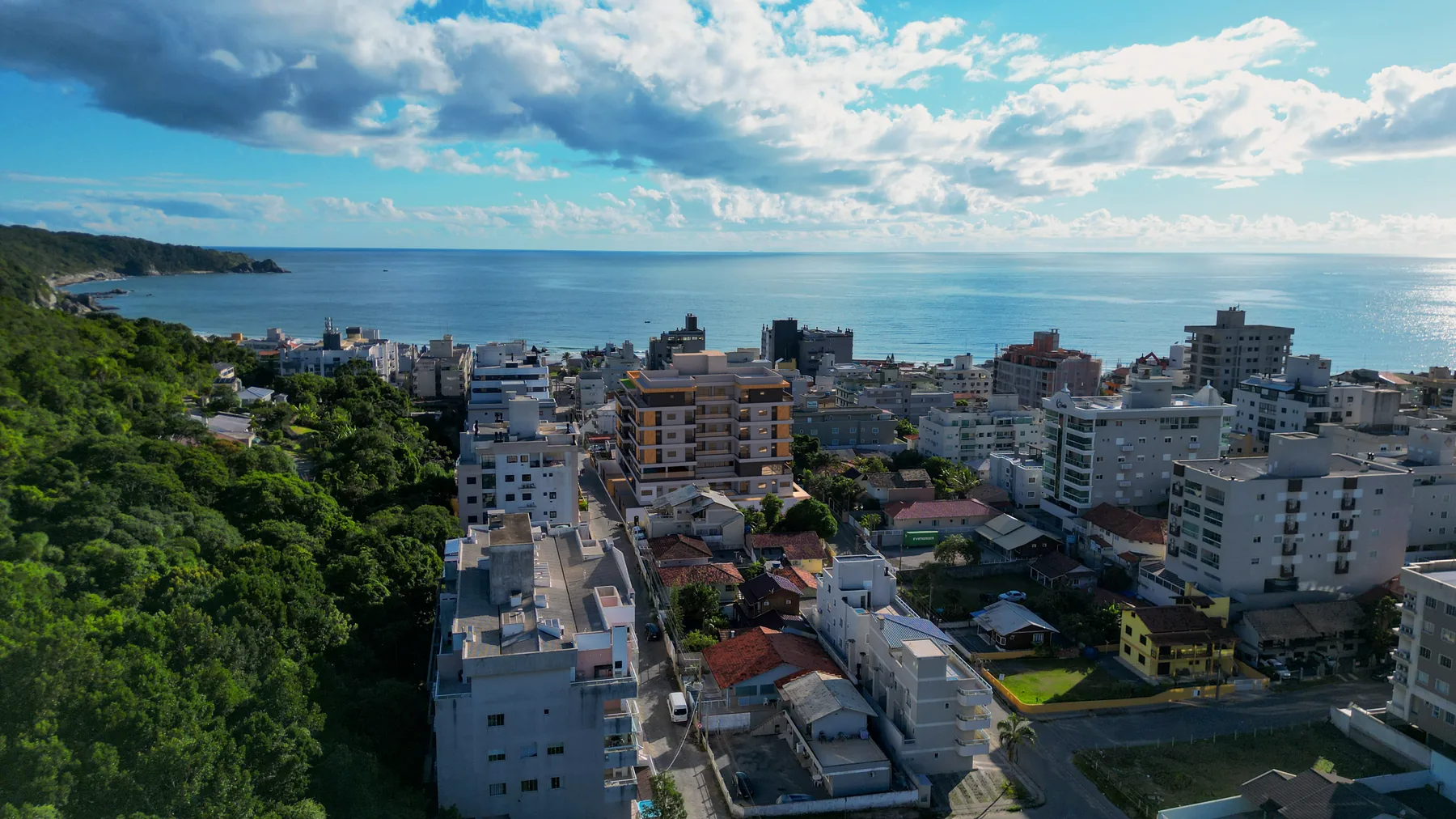Vista aérea do bairro de Bombas a partir do morro, com a mata em primeiro plano, as casas e prédios baixos e o mar aberto ao fundo