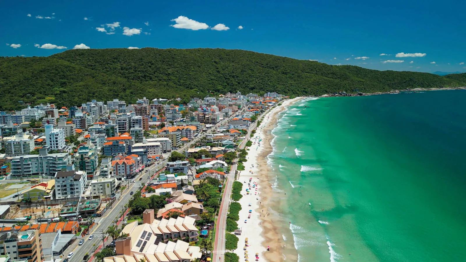 Vista aérea de uma enseada da região, com o casario entre o morro e a faixa de areia e o mar esverdeado