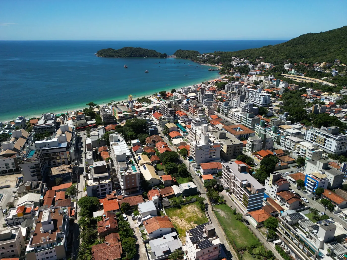 Foto aérea do Centro de Bombinhas, com a faixa de areia à esquerda, a enseada de água clara, o morro coberto de mata ao fundo e o miolo de casas e prédios baixos à frente