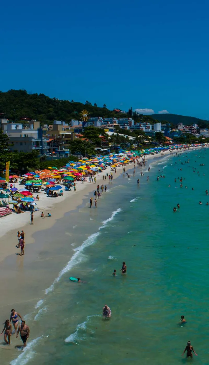 A praia do Centro de Bombinhas vista de cima, com guarda-sóis coloridos na areia, banhistas na água rasa e o morro verde na ponta
