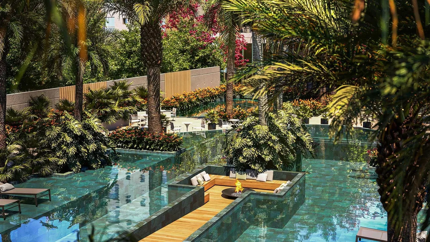 Piscina alongada cercada de palmeiras e vegetação, com deck de madeira e as torres ao fundo