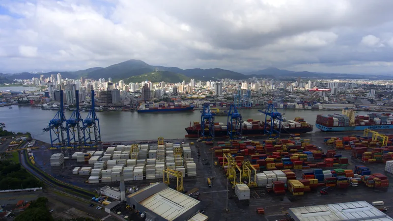 Vista aérea do porto de Itajaí, com os guindastes e os contêineres no cais e a cidade do outro lado do canal