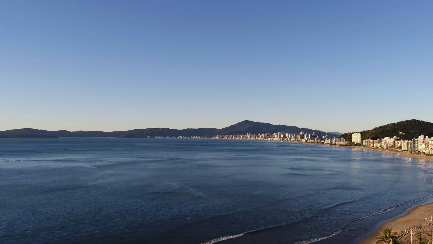 Vista aérea da orla de Itapema ao amanhecer, com a faixa de areia curva, o mar calmo e os morros ao fundo