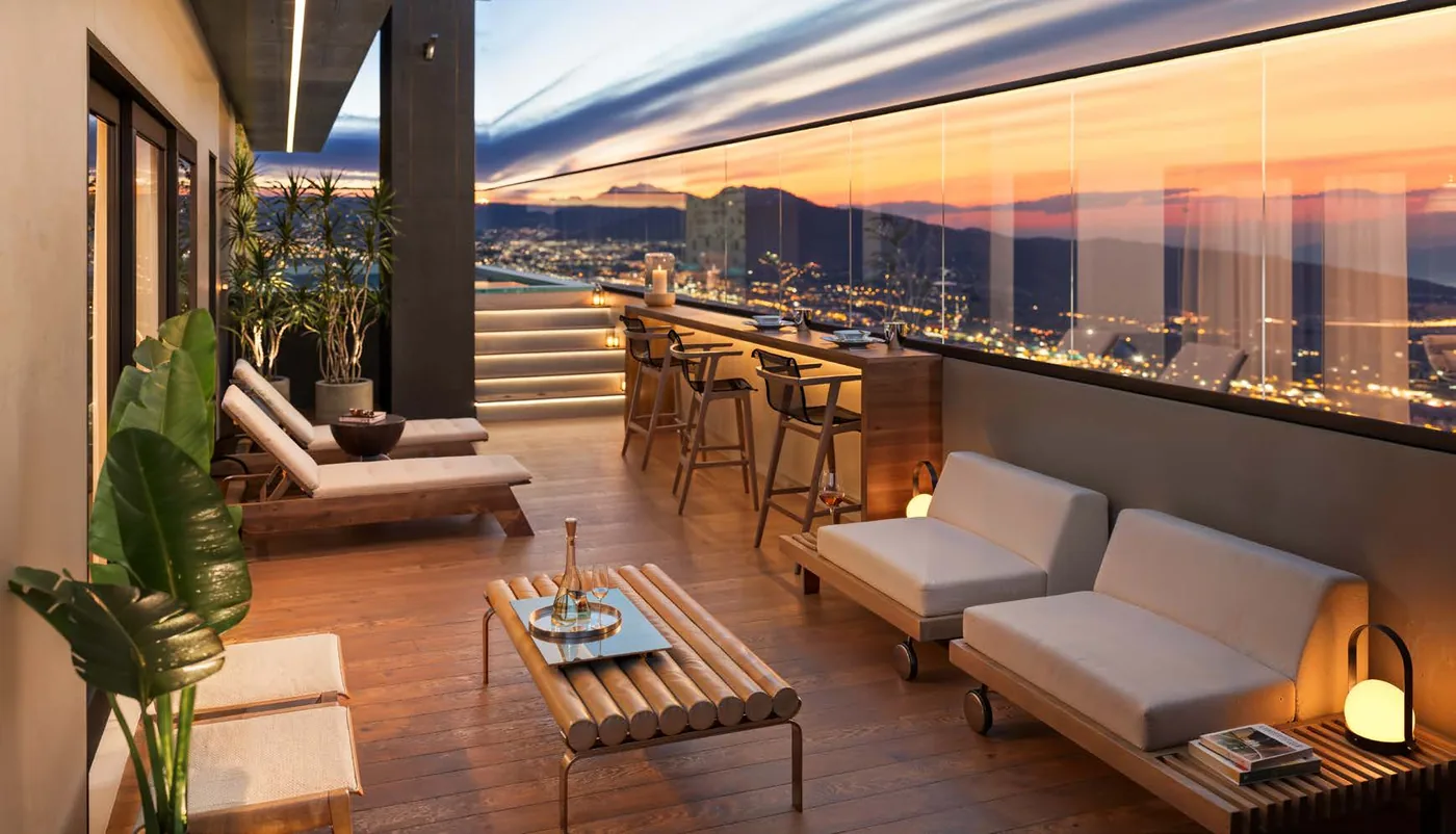 Solarium do rooftop ao pôr do sol, com espreguiçadeiras, bancada alta junto ao peitoril de vidro e a cidade ao fundo