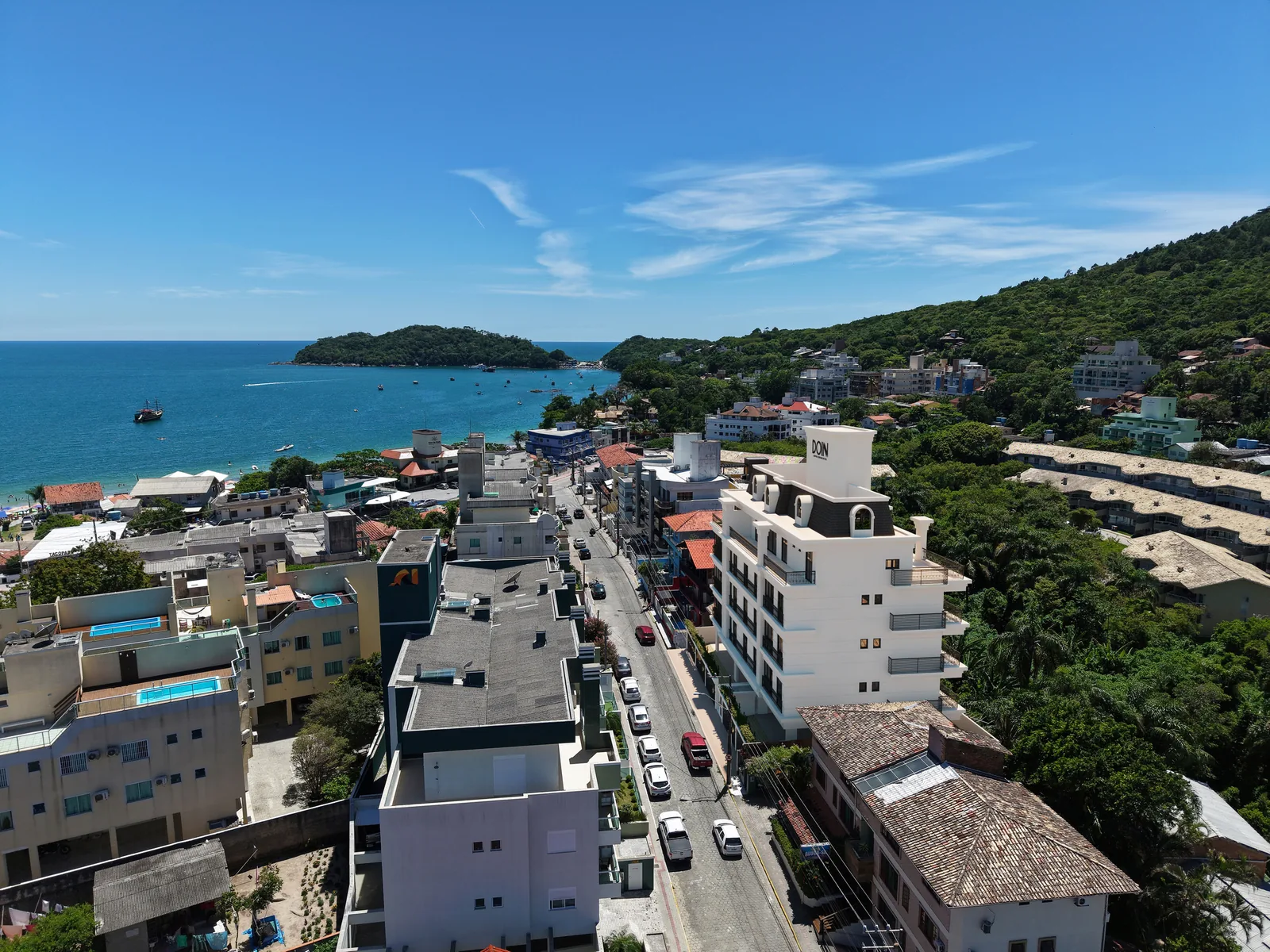 Vista aérea da rua do Château de Lumière descendo até o mar, com a enseada, a ilha e o costão ao fundo, e o prédio inserido entre as casas à direita