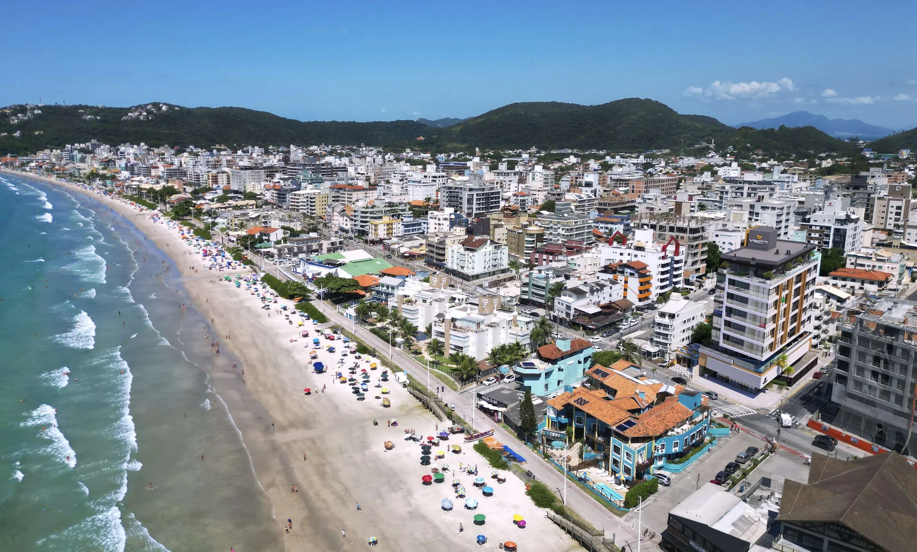 Vista aérea da Praia de Bombas, com a faixa de areia cheia de guarda-sóis, o mar verde à esquerda, a cidade baixa à direita e os morros ao fundo