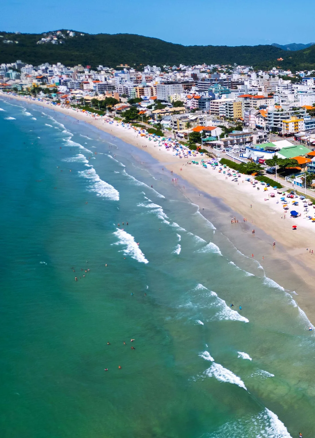 A orla de Bombas vista de cima em enquadramento vertical, com a arrebentação, a areia clara e a cidade acompanhando a curva da praia