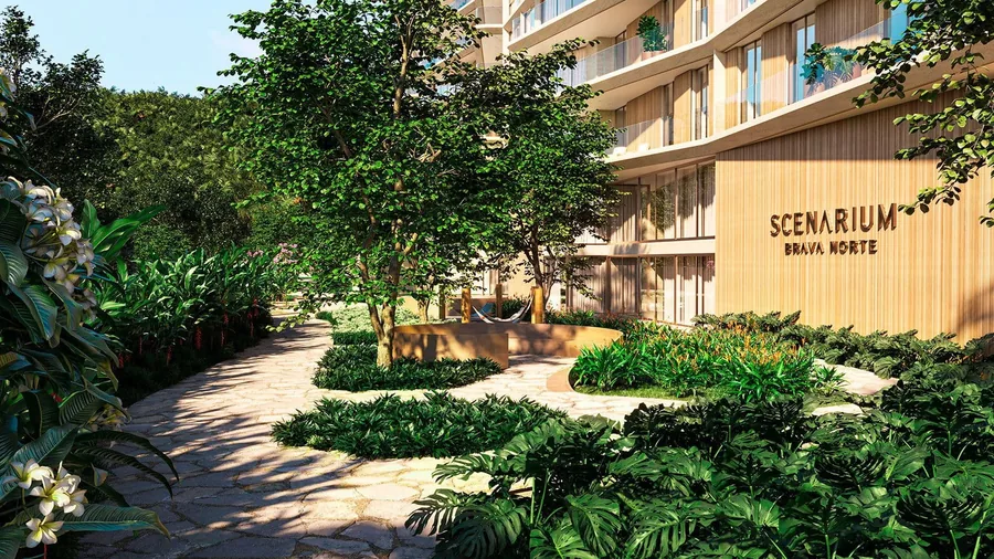 Acesso ao Scenarium entre jardins densos, com o letreiro do empreendimento em muro claro sob as árvores