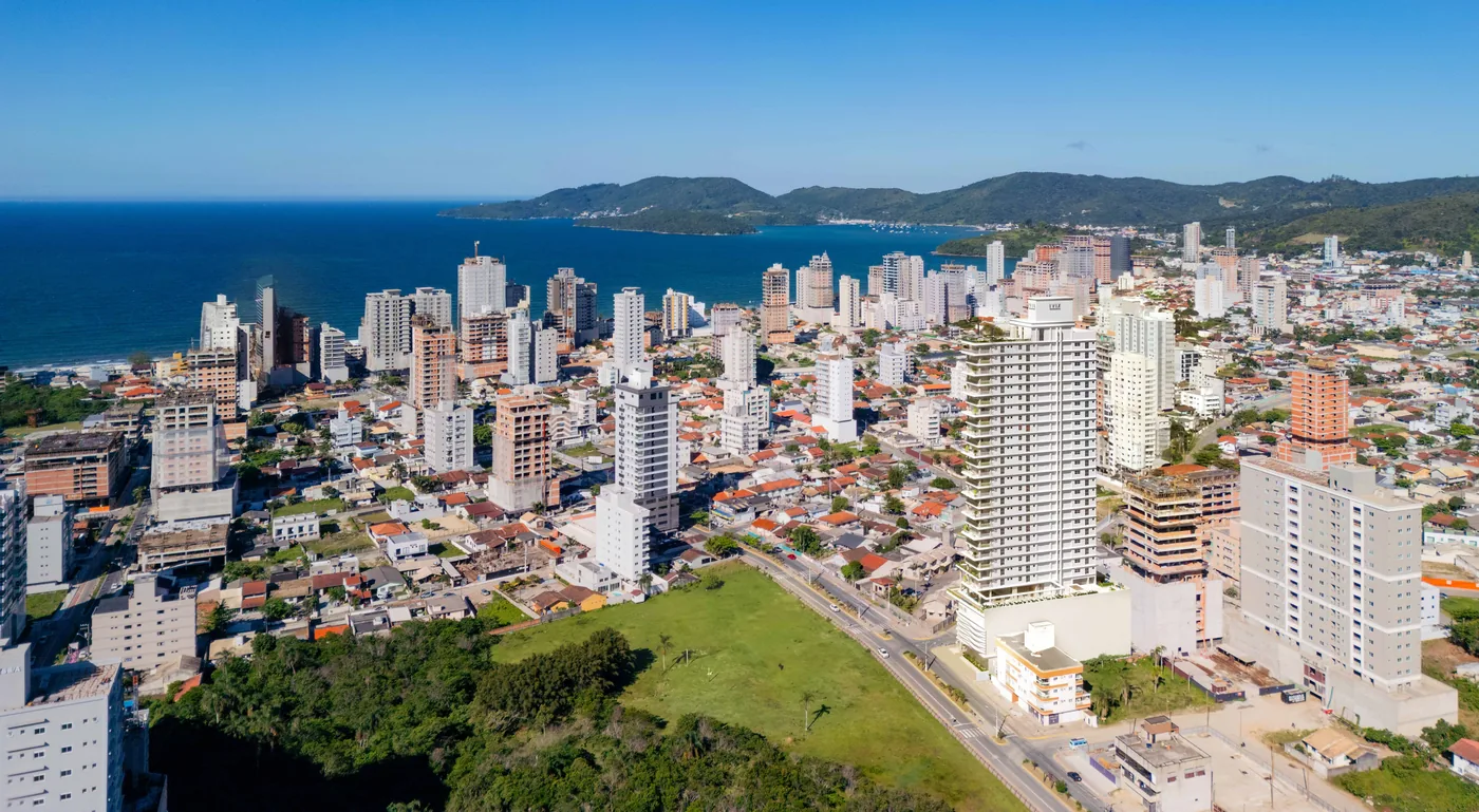 Vista aérea de Balneário Perequê, em Porto Belo, com a malha de casas e prédios baixos, a orla e os morros da costa ao fundo