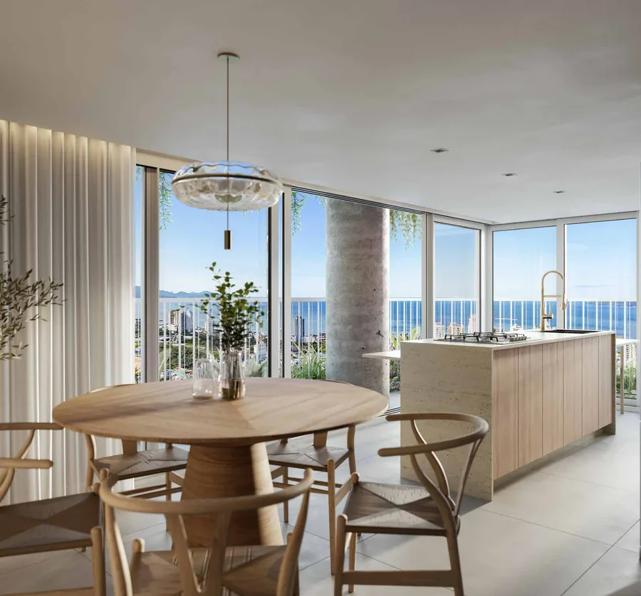Canto de jantar do apartamento, com mesa redonda de madeira, cadeiras de palha, pendente de vidro e a cozinha clara atrás